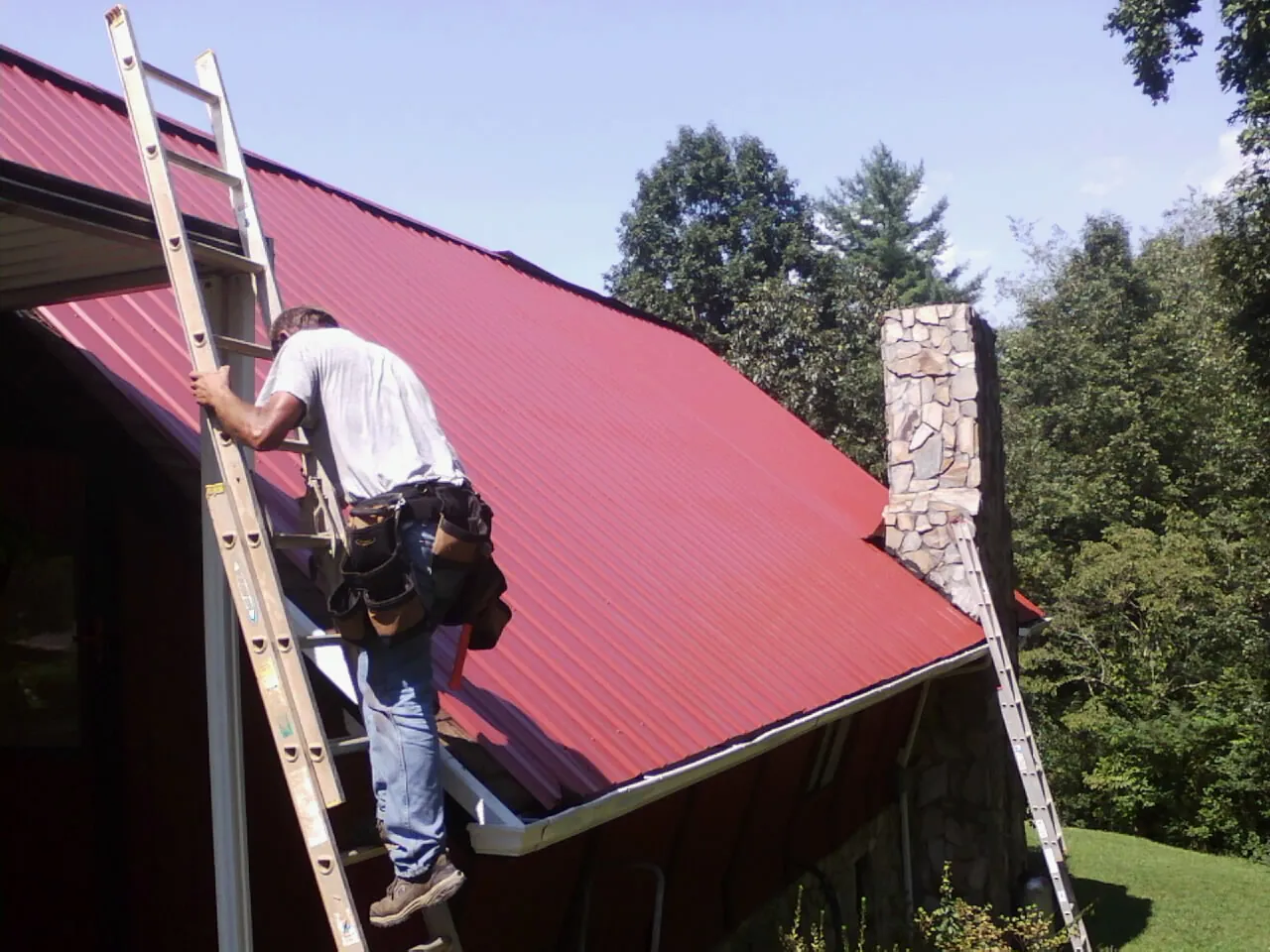 Roofer on a ladder beside a red metal roof and stone chimney — Soffit & Fascia Repair by Roofing Brothers in Aaronsburg